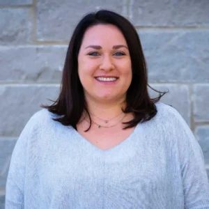 Woman with dark shoulder-length hair wearing light gray v-neck top, smiling at camera against brick wall