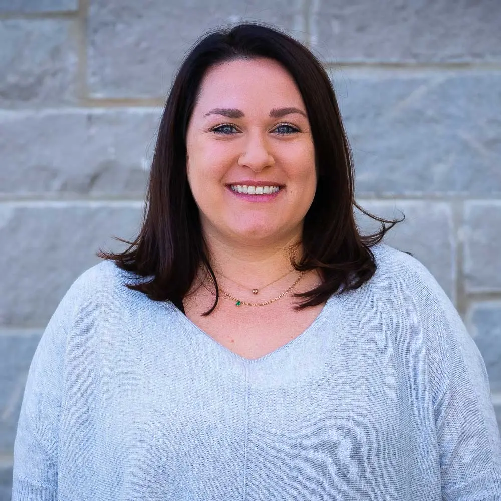 Woman with dark shoulder-length hair wearing light gray v-neck top, smiling at camera against brick wall