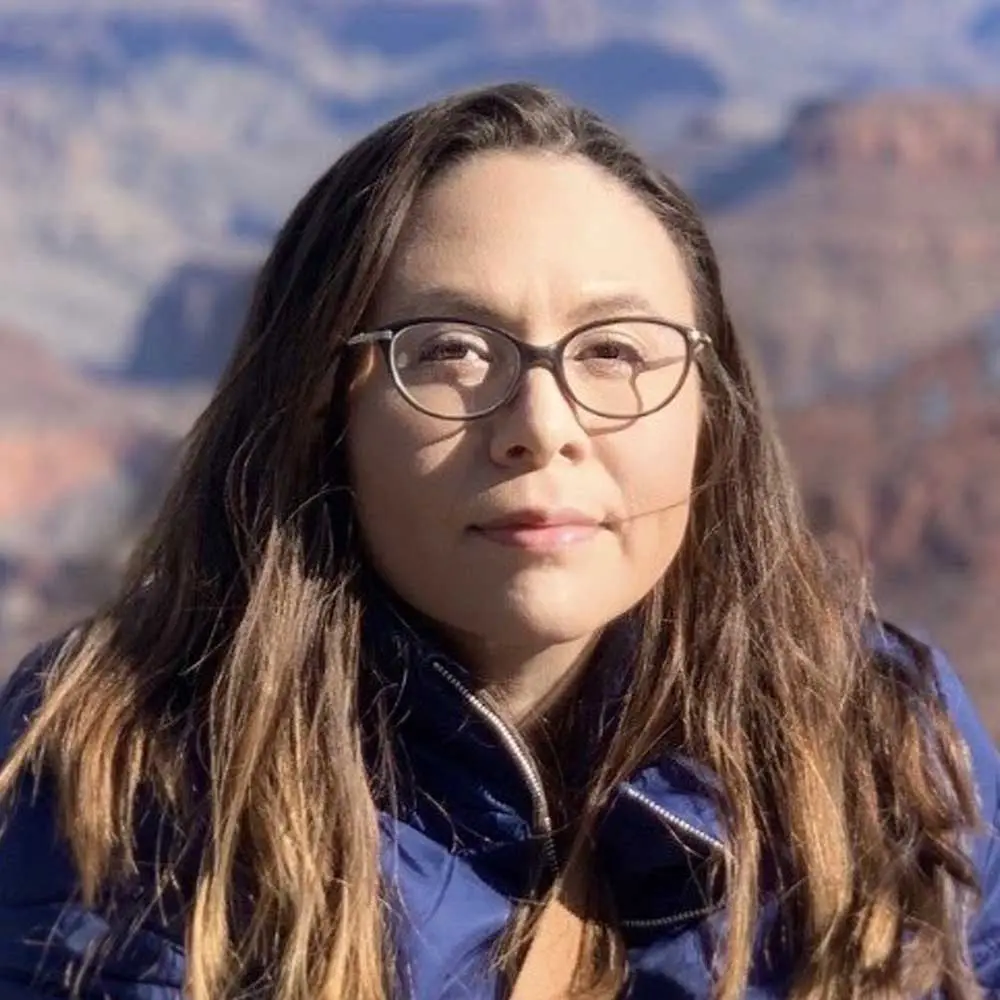 Woman with long brown hair and glasses wearing a dark blue jacket, looking at camera with a scenic mountainous background.