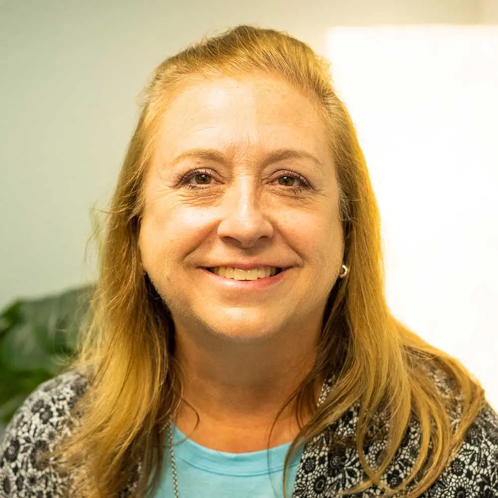 Woman with reddish-blonde hair pulled back, smiling at camera indoors, wearing blue shirt and patterned cardigan