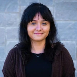 A portrait of a woman with long dark hair and bangs wearing a dark cardigan over a black shirt, smiling outdoors