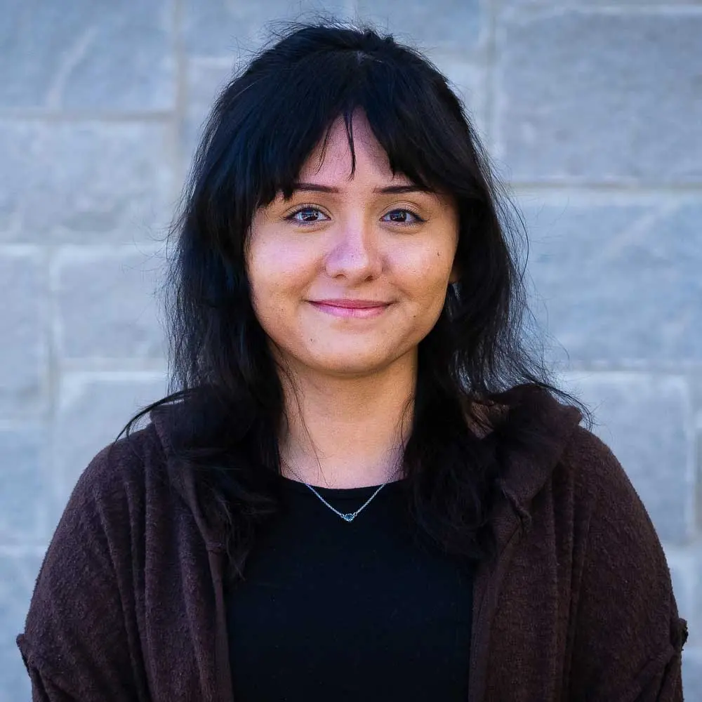 A portrait of a woman with long dark hair and bangs wearing a dark cardigan over a black shirt, smiling outdoors