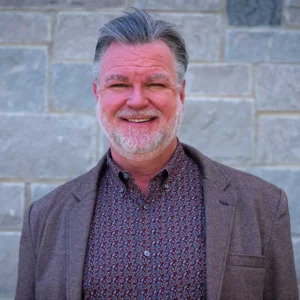Professional headshot of a middle-aged man with gray hair and beard, wearing a brown blazer over a patterned purple shirt, smiling against a brick ...