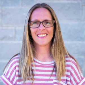 Woman with long brown hair and glasses wearing pink and white striped shirt, smiling at camera against blurred background