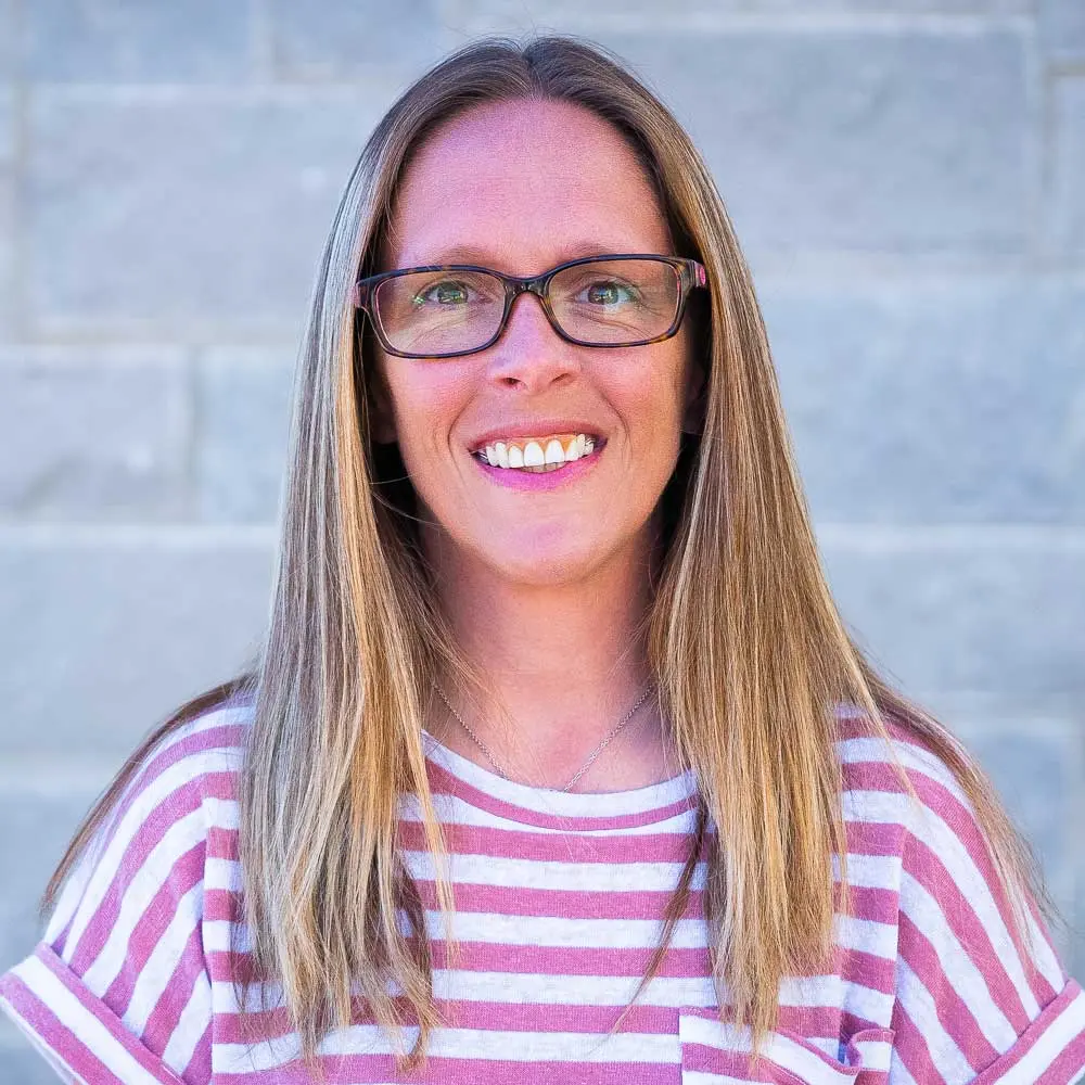 Woman with long brown hair and glasses wearing pink and white striped shirt, smiling at camera against blurred background