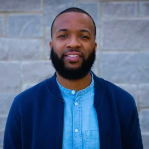 Man with dark skin wearing light blue button-up shirt under dark blue cardigan, smiling at camera against brick wall