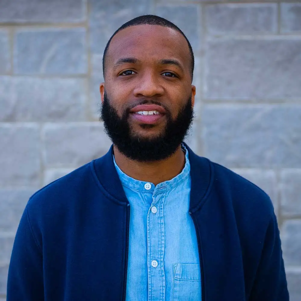 Man with dark skin wearing light blue button-up shirt under dark blue cardigan, smiling at camera against brick wall