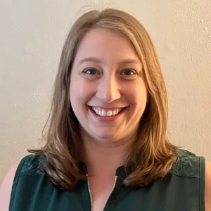 Woman with shoulder-length reddish-brown hair wearing dark green top, smiling at camera against beige wall