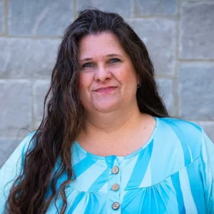Woman with long dark hair wearing a turquoise and white patterned top, smiling at the camera against a textured gray stone background.