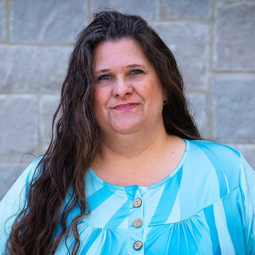 Woman with long dark hair wearing a turquoise and white patterned top, smiling at the camera against a textured gray stone background.