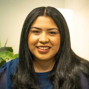 Woman with long dark hair wearing a dark navy top, smiling at camera with a potted plant visible behind her shoulder.