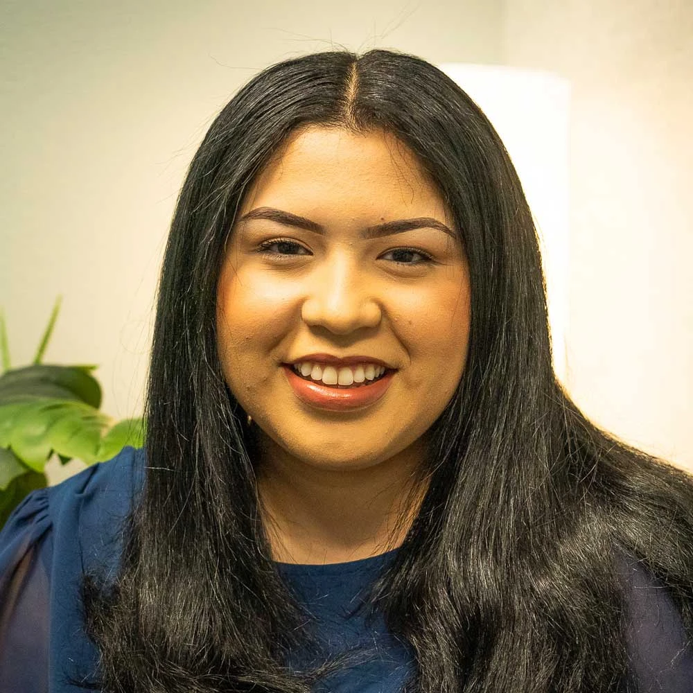 Woman with long dark hair wearing a dark navy top, smiling at camera with a potted plant visible behind her shoulder.