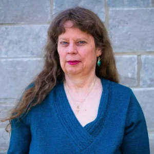 Professional headshot of a woman with long brown wavy hair wearing a blue v-neck sweater and teal earrings against a brick wall background.