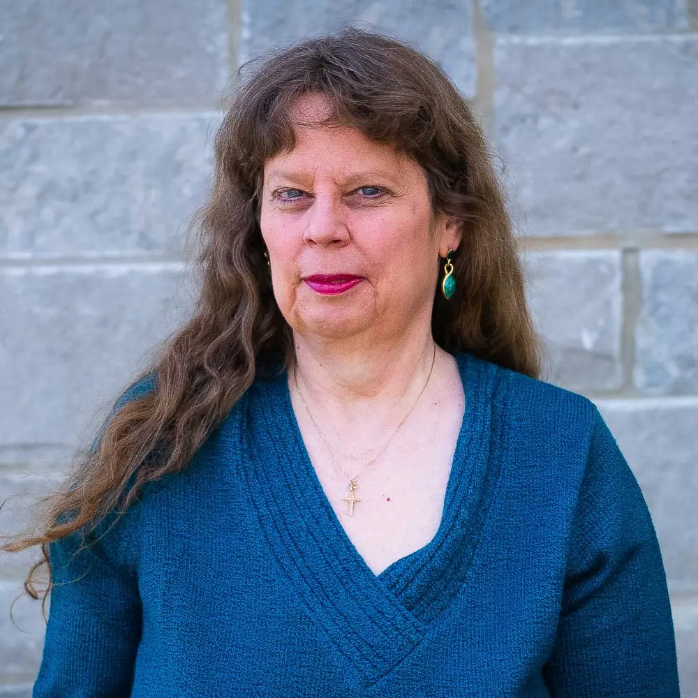 Professional headshot of a woman with long brown wavy hair wearing a blue v-neck sweater and teal earrings against a brick wall background.