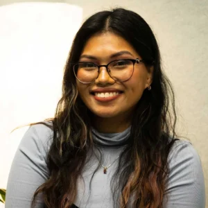 Young woman with long black hair and glasses, wearing a gray shirt, smiling at the camera in an indoor setting with white walls.