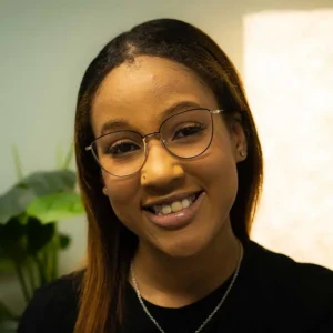 Young woman with long black hair and glasses, smiling at the camera while wearing a black top in front of a blurred green plant background.