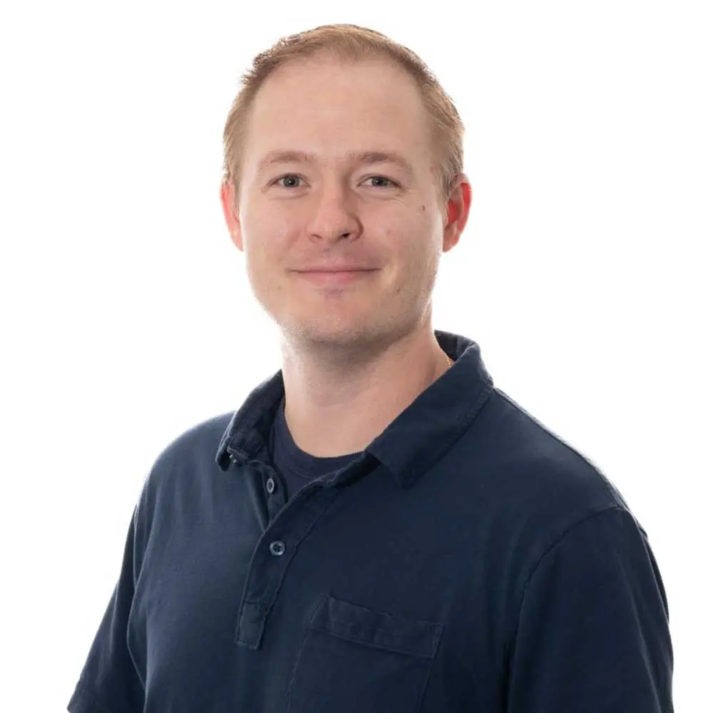A professional headshot of a man with reddish-blonde hair wearing a dark blue collared shirt, smiling at the camera
