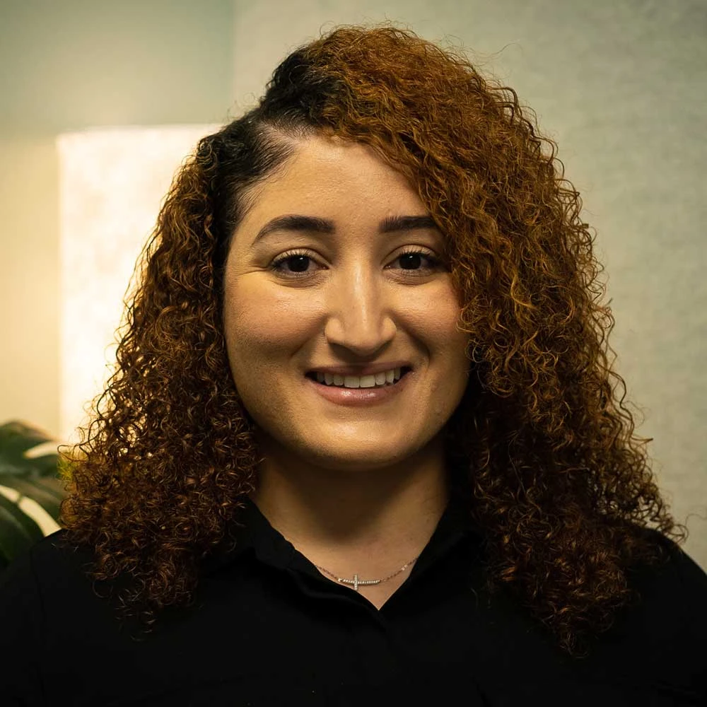 Woman with long curly reddish-brown hair wearing a black top, smiling warmly at camera against neutral background