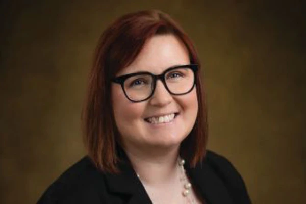 Woman with reddish-brown hair and glasses wearing gray patterned top smiling at camera while seated indoors against brick wall background