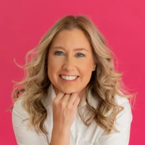 Woman with blonde wavy hair smiling at camera against pink background wearing white collared shirt